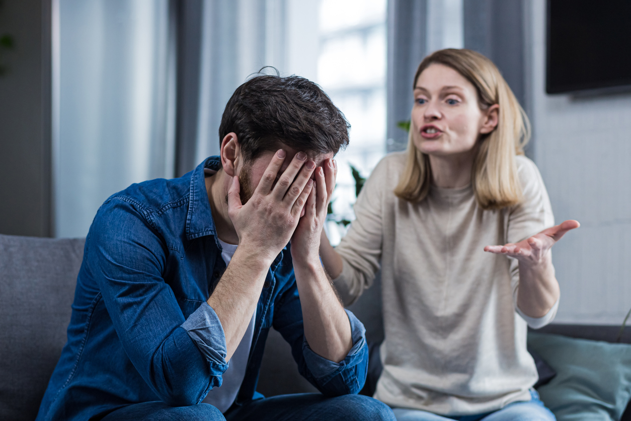 A couple having an argument while a man in a blue shirt puts his face in his hands and his partner yells at him.