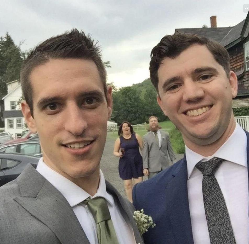 Two guys try to pose for wedding photo but the man and woman behind them crash the photo by making silly faces. 
