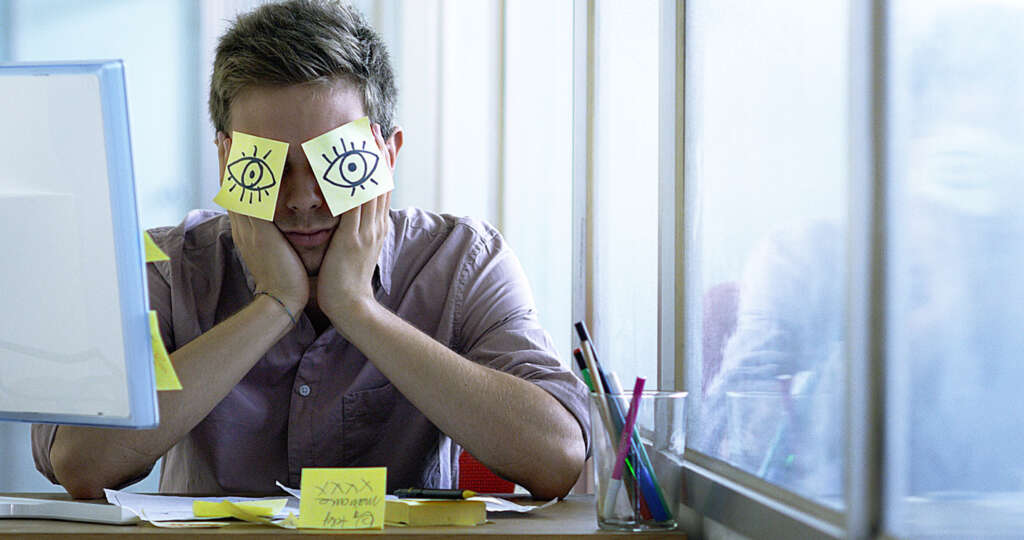 A young employee who fell asleep at his work desk in front of his large monitor and now has sticky notes covering his eyes. 