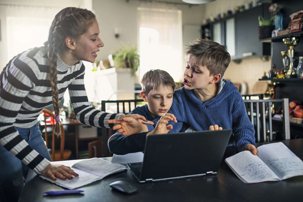 Mother angrily trying to keep her two sons calm while one of them sits in front of the computer doing homework. 