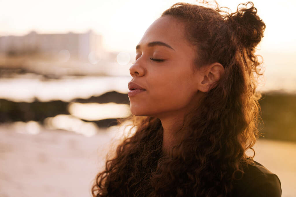 Woman sitting in the sunshine with her eyes closed, relaxing, and breathing outward. 