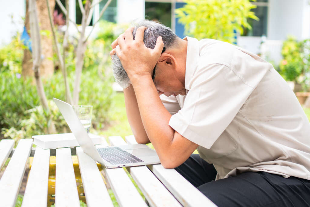 Man sitting at outdoor table in front of his laptop with his head angrily in his hands. 