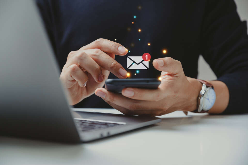 A person with a nice watch on sending an email out from their phone while sitting in front of their laptop. 