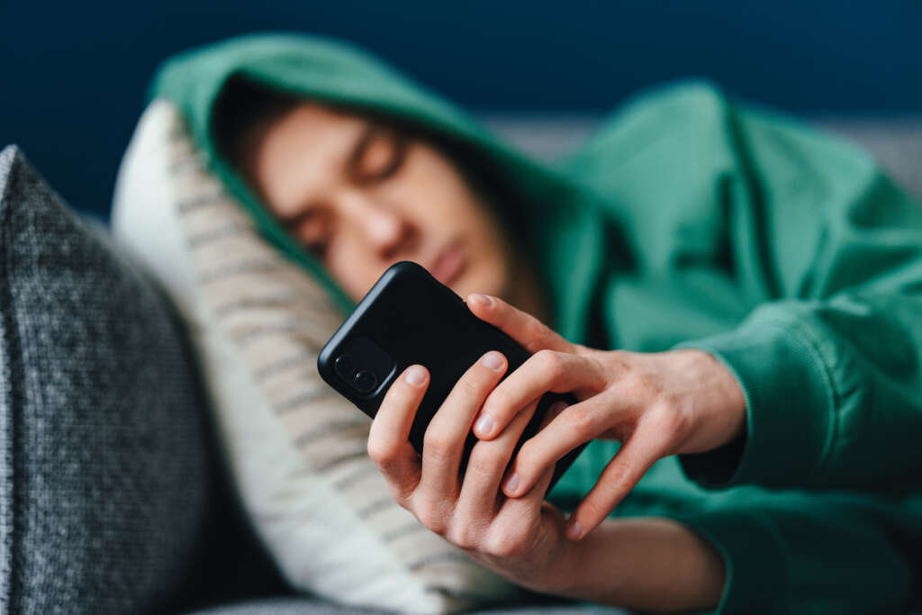 Kid in green sweatshirt with hood on keeping busy using their phone,. 