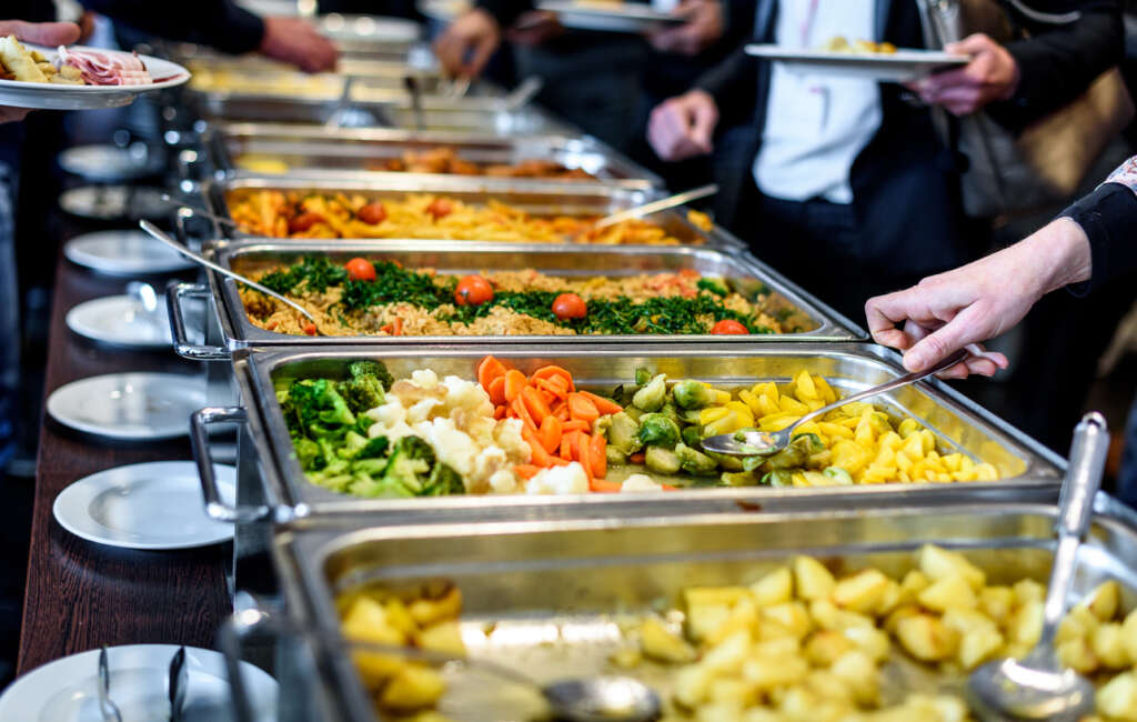 Employees that are taking various food items from a work cafeteria's lunch buffet. 