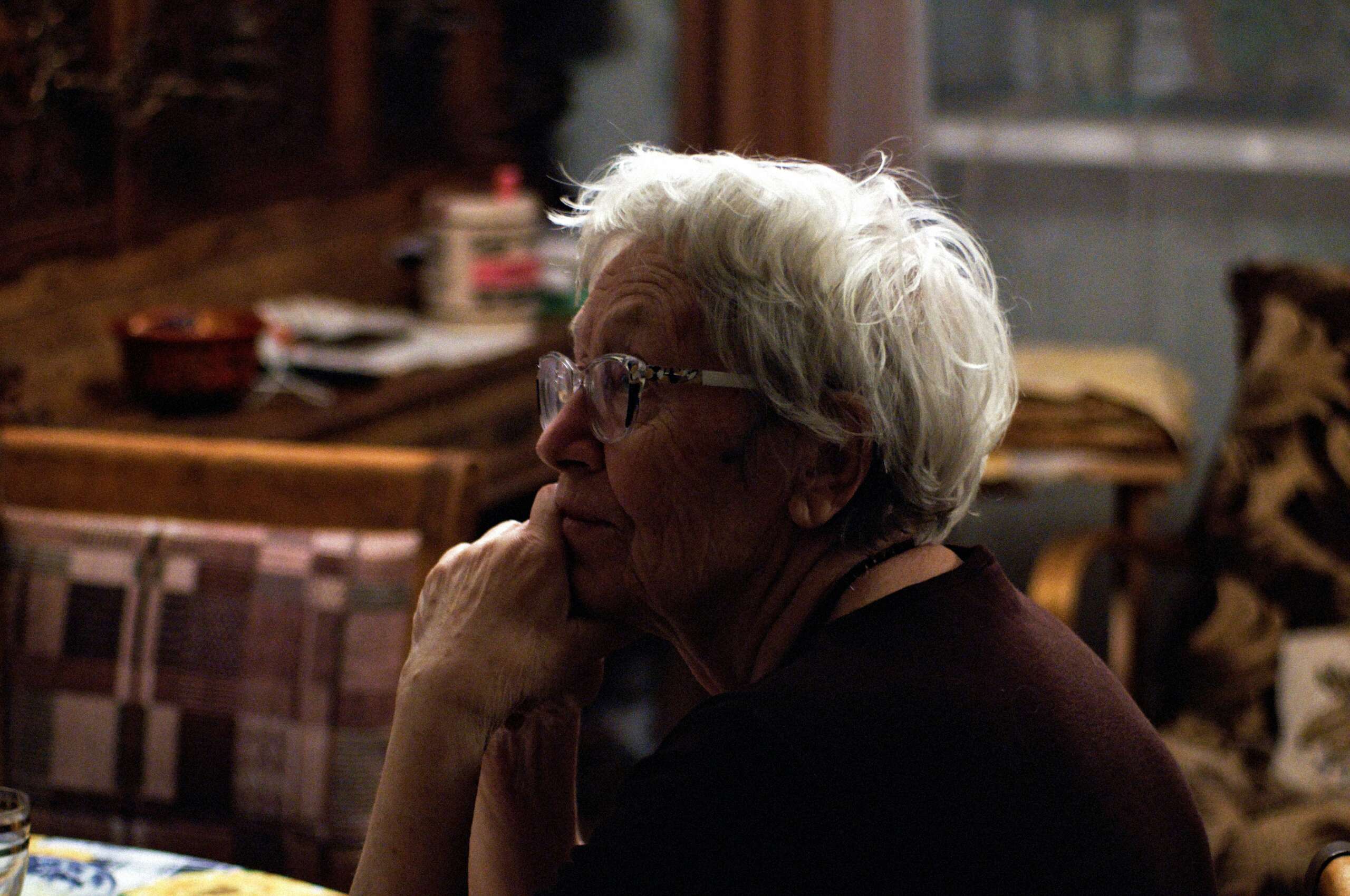 An image of an elderly woman sitting at a table with her face in her hands.