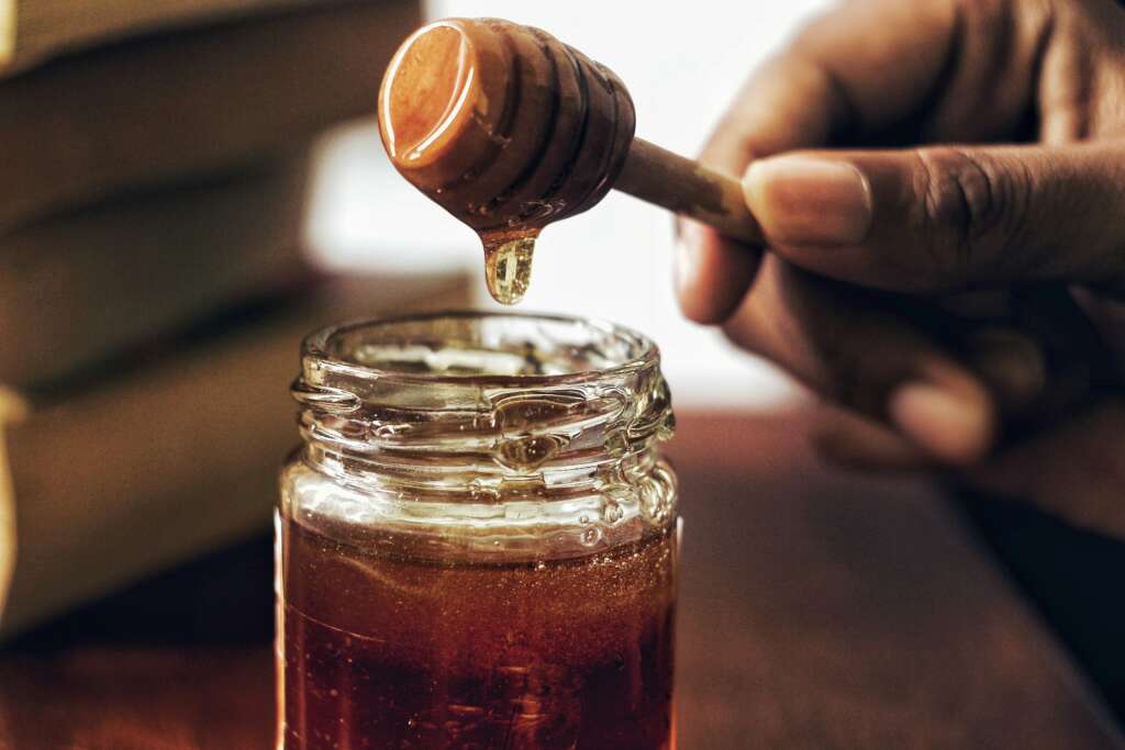 A person using a honey dipping tool to grab honey from a honey jar. 