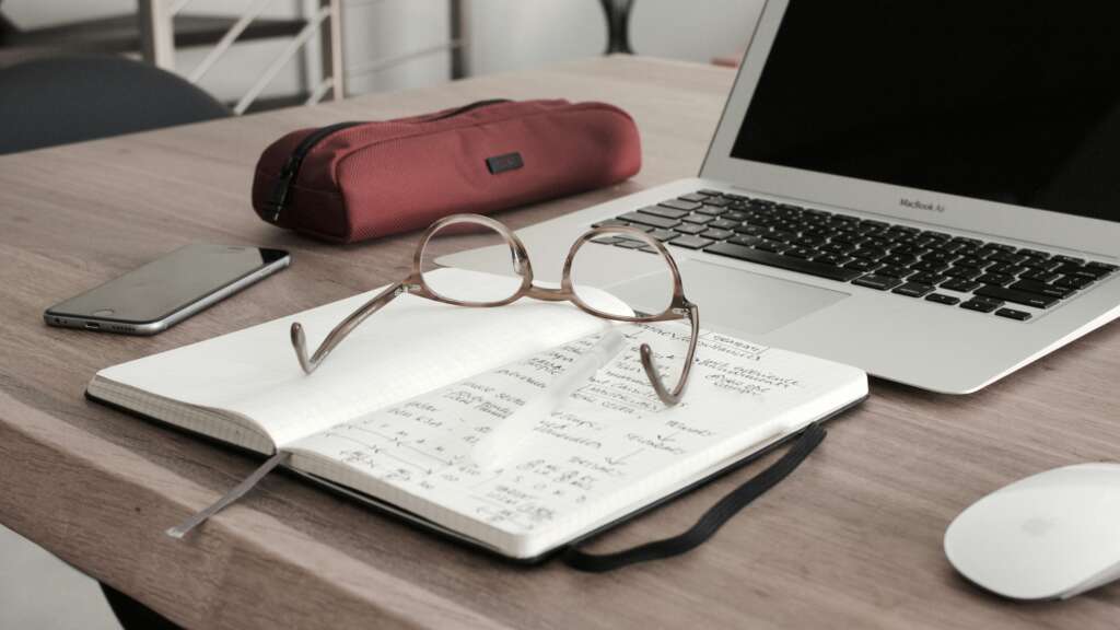 An image of glasses on a notebook in front of a laptop with a mouse placed next to it. 