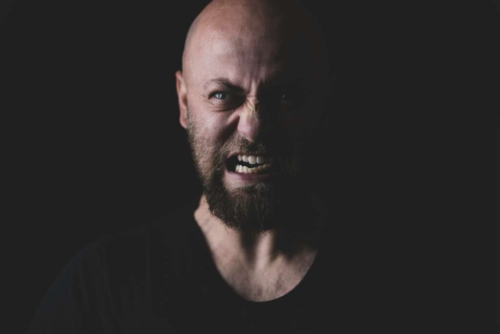 A very angry man with a beard wearing a black shirt with a black background. 