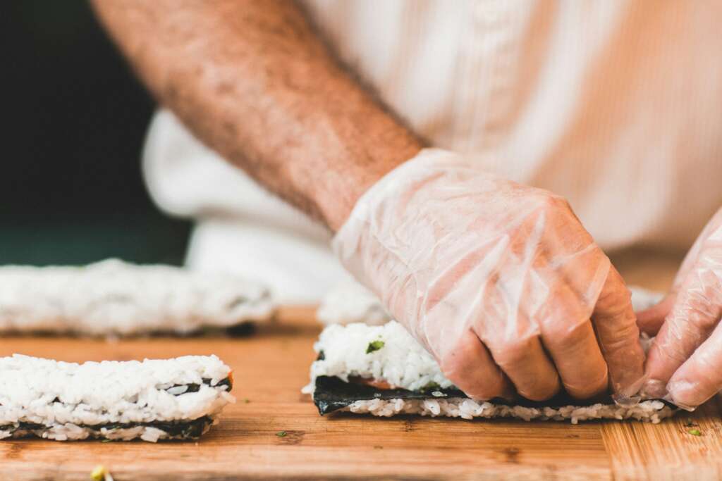 An image of a person with cooking gloves on preparing a sushi roll. 