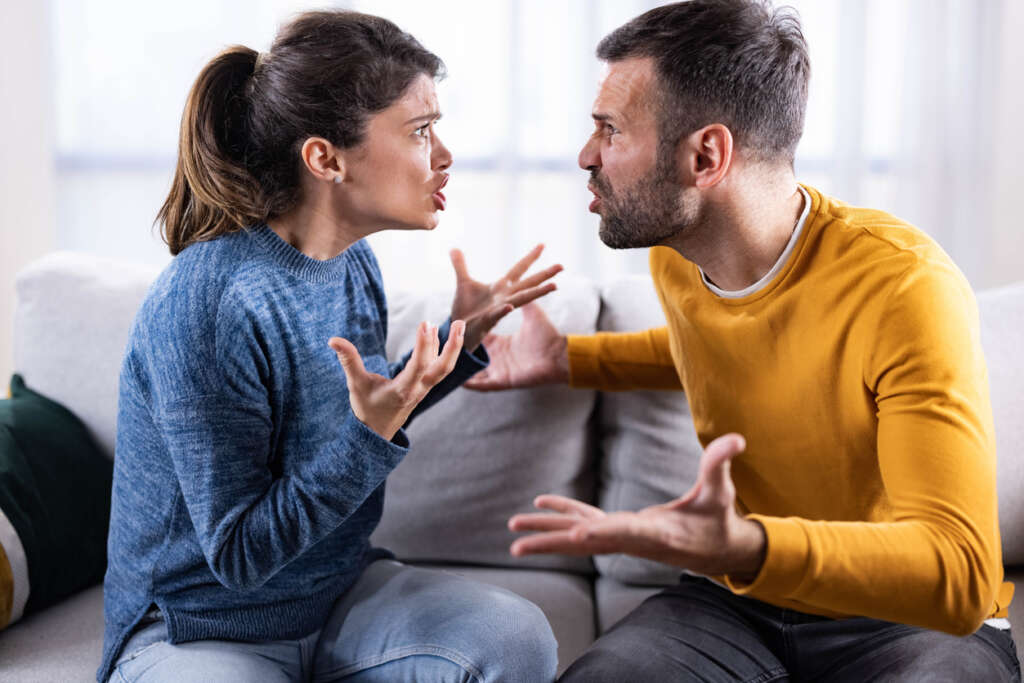 A couple having an argument on their couch while one person wears a yellow sweater and the other person wears a blue sweater. 