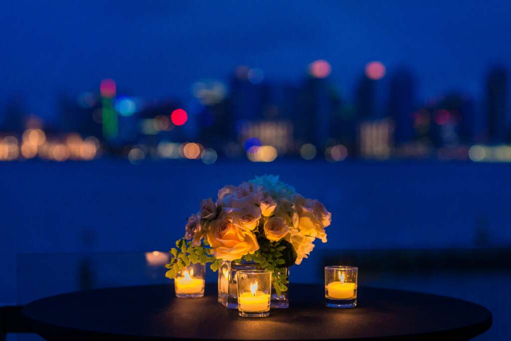 A candlelit dinner table with a vase of flowers and a nice view of the city at night. 