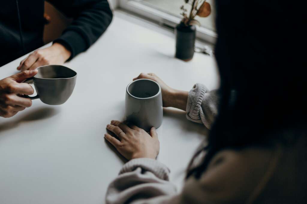 An image of two people with coffee mugs sitting at a table. 