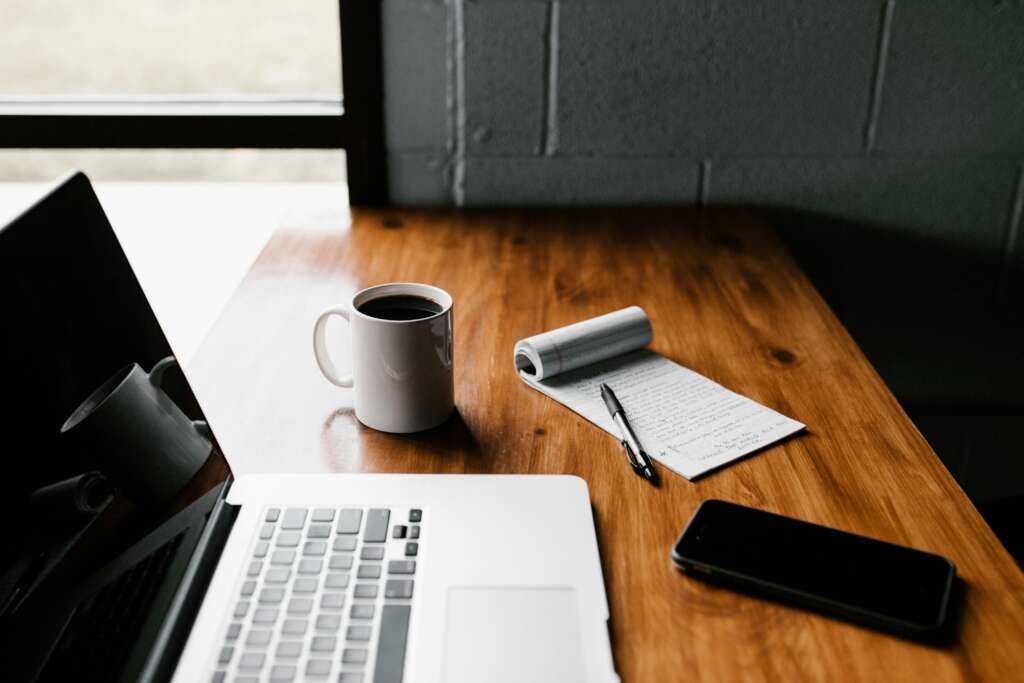 An image of a notepad placed next to a phone, a mug of coffee, and a laptop. 