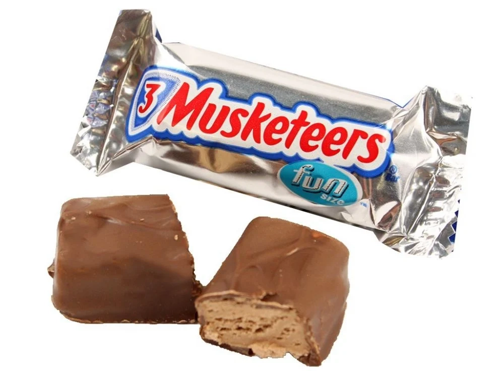 A 3 Musketeers fun size candy bar in a silver wrapper is displayed. In front of the wrapper, the chocolate bar is cut in half, showing its creamy nougat filling.