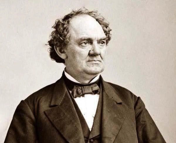 A sepia-toned portrait of a middle-aged man with curly hair. He is wearing a dark suit, a bow tie, and a white shirt. The man has a stern expression and is looking slightly to the right. The background is plain and unobtrusive.