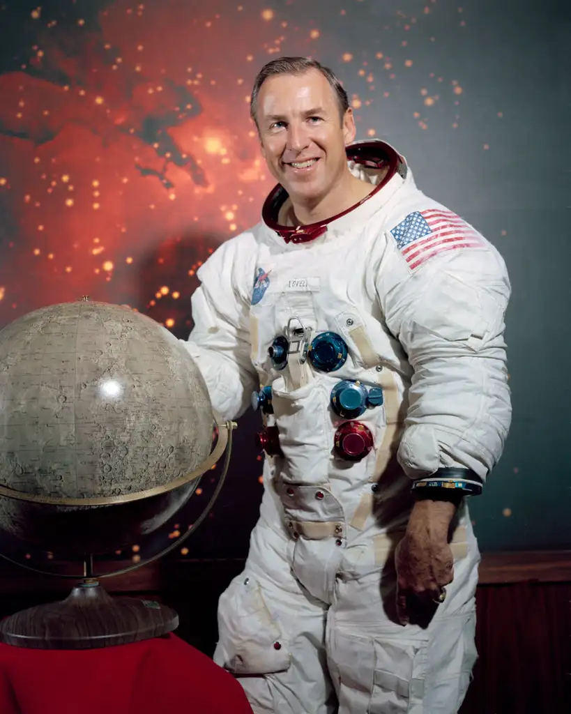 A smiling astronaut in a white spacesuit with an American flag patch stands indoors next to a moon globe. The background features a starry space scene with red hues. The astronaut has short, neat hair and poses with one hand on the globe.