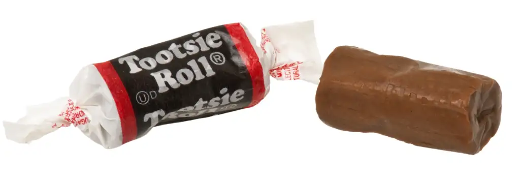 A Tootsie Roll candy is shown. One piece is still wrapped in its black, white, and red wrapper with the "Tootsie Roll" logo visible, while another piece, unwrapped, reveals its cylindrical, elongated chocolate-flavored taffy-like appearance.