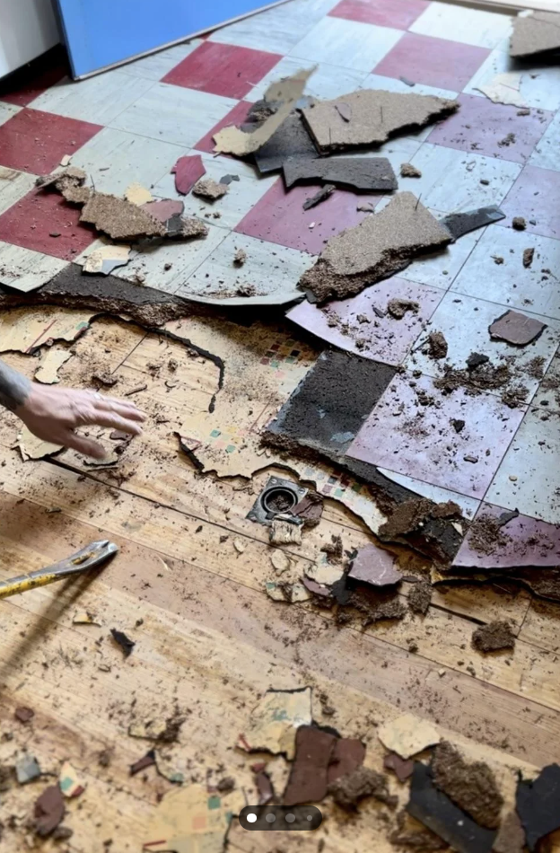 A partially removed vinyl tile floor reveals a deteriorating and decayed subfloor beneath. Pieces of red, gray, and cream-colored tiles are scattered around. A hand with a tool is visible on the left side, actively working on removing the tiles.