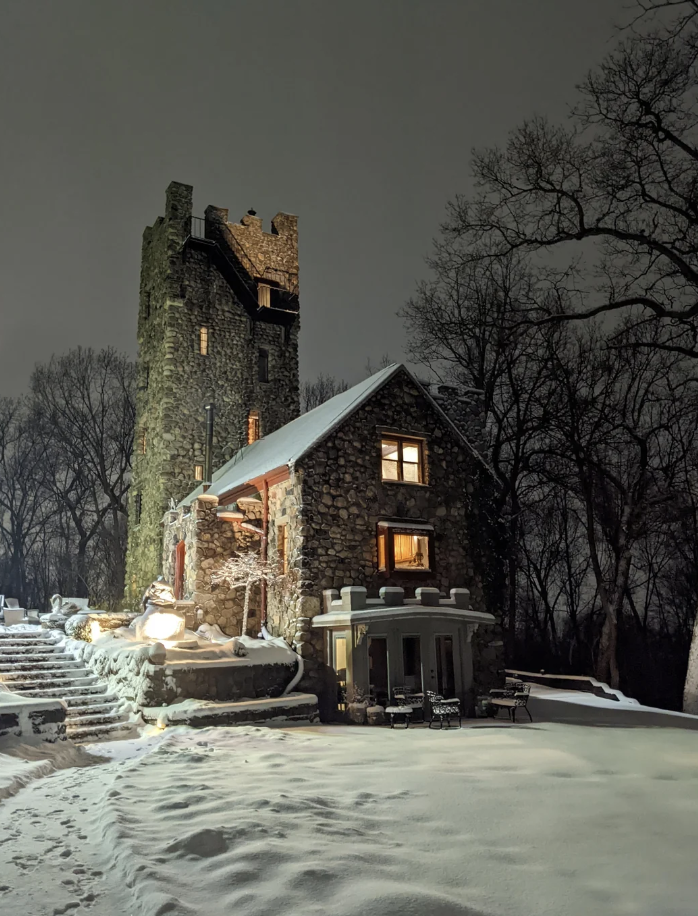 A stone castle-like house surrounded by snow, illuminated under a night sky. There are lights on inside the house, casting a warm glow through the windows. Bare trees frame the background, and snow-covered steps lead up to the entrance.
