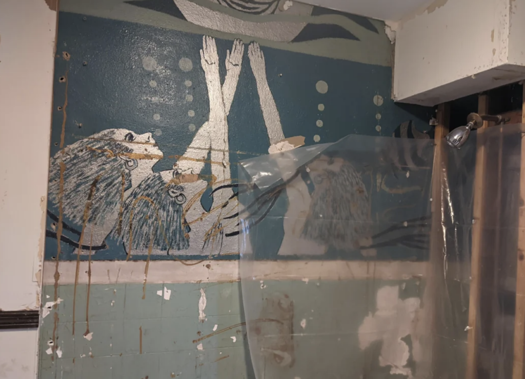 A mural on a bathroom wall depicts several abstract figures with wavy hair and reaching arms, surrounded by bubbles. The bathroom is under renovation, with exposed pipes and a clear plastic sheet hanging nearby. The mural is in shades of blue, white, and gray.