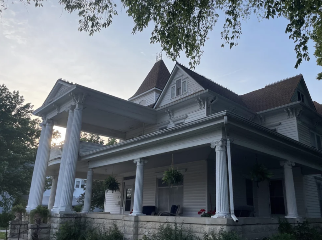 A large, historic white house with a wrap-around porch and classical columns. The house has a gabled roof and an upper balcony. Hanging plants and greenery decorate the porch area. The sky is clear with the sun setting or rising behind the house. Trees surround the area.