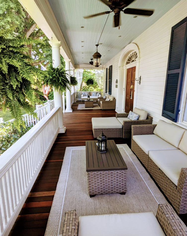 A spacious, wooden-floored porch with white railings features comfortable wicker furniture with cream cushions, a ceiling fan, hanging plants, and a lantern on a coffee table. The porch is surrounded by greenery and leads to a door with an arched window.