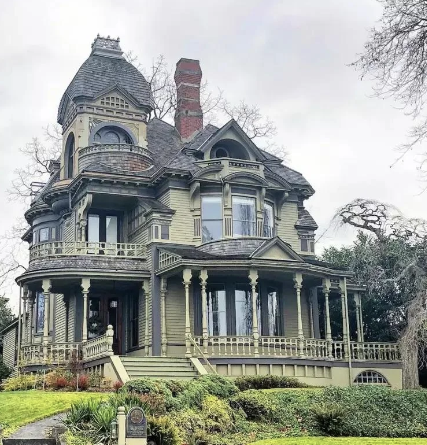 A large, Victorian-style house with a round turret, ornate detailing, and multiple chimneys. The home features balconies, numerous windows, and is painted in muted green and brown tones. It is surrounded by a partially visible garden with shrubs and flowering plants.