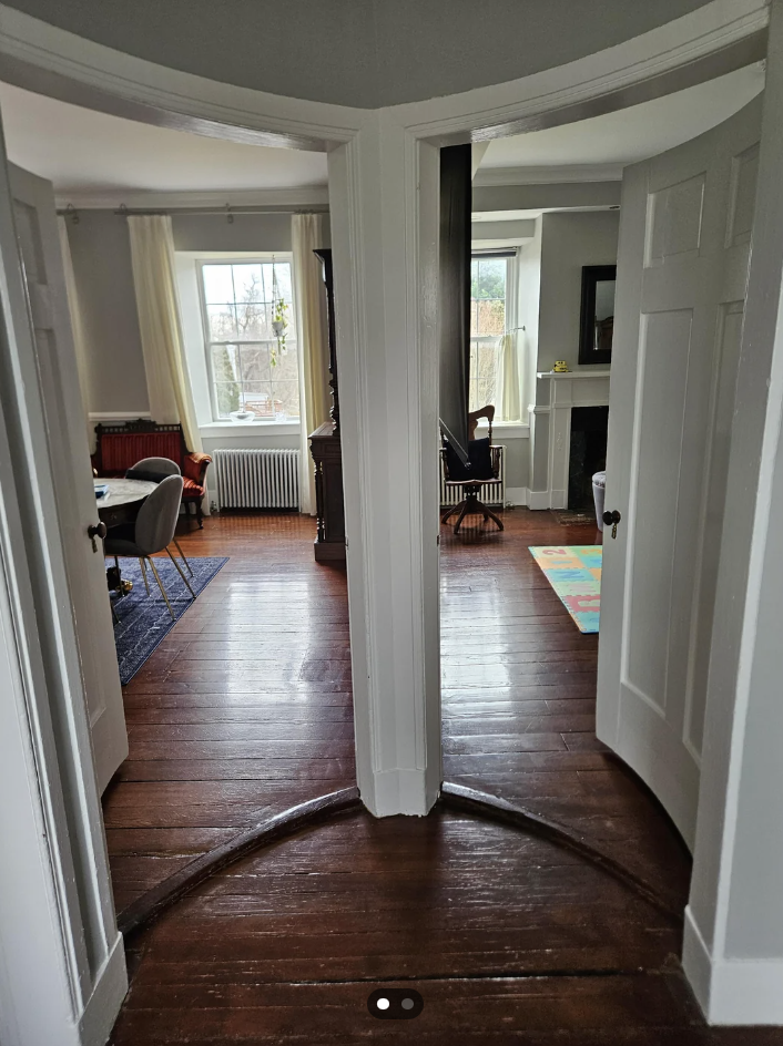 A split-door entryway opens into two bright rooms with white walls and wooden floors. The room on the left has a red sofa and a round table, while the room on the right has a fireplace, a window with curtains, and a colorful rug. Both rooms have large windows.