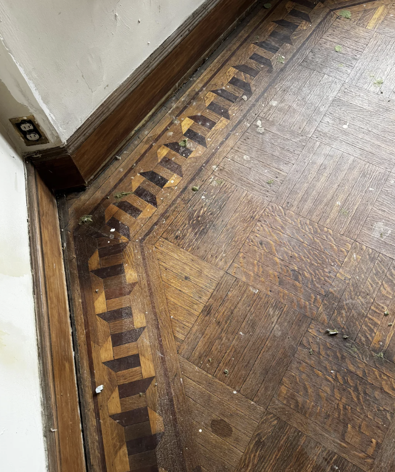 A close-up view of a corner in a room showcasing a wooden parquet floor with intricate geometric patterns near the edge. The floor has various shades of brown and some debris scattered on it. The base of the wall is visible with a wooden skirting and an electrical outlet.