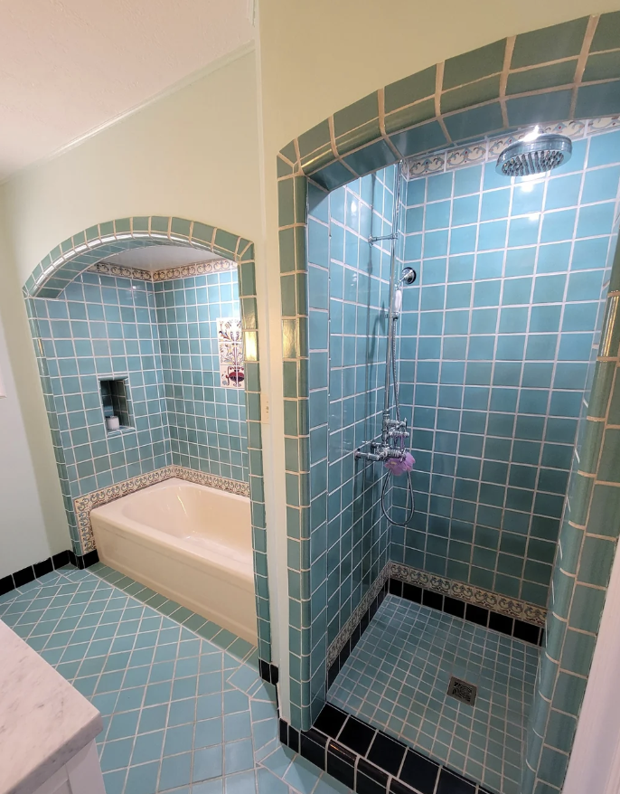A bathroom with teal tiles features a walk-in shower on the right and a bathtub on the left. Both the shower and bathtub are framed by arched openings. The floor is covered with matching teal tiles bordered with black trim.