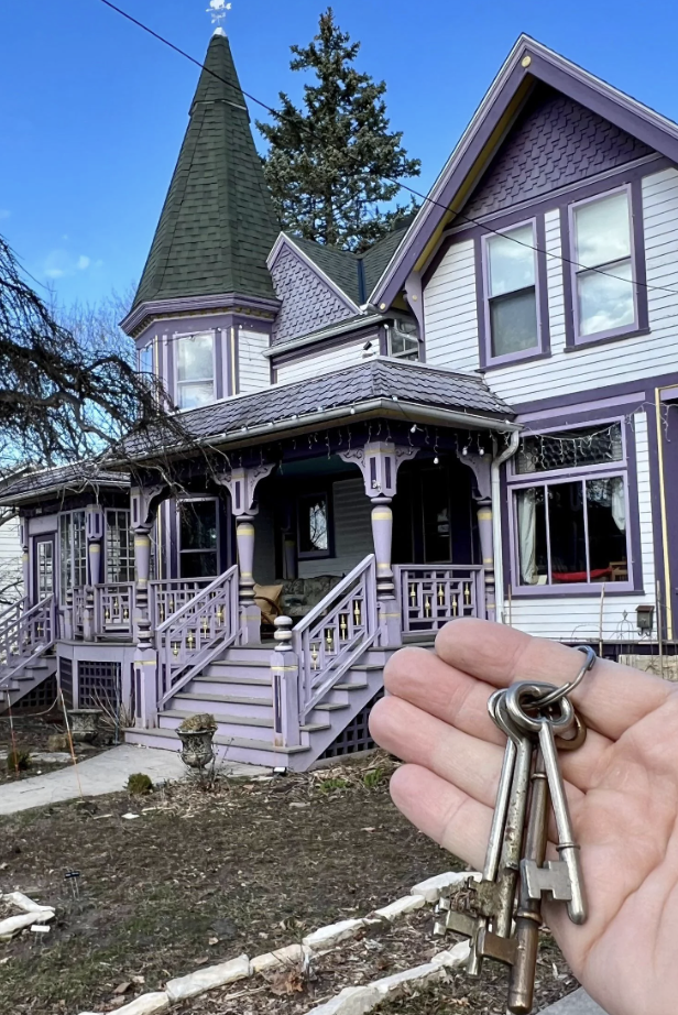 A hand holding vintage keys in the foreground, with a large, ornate Victorian-style house painted in lavender and purple visible in the background. The house features pointed roofs, ornate woodwork, and a wraparound porch.