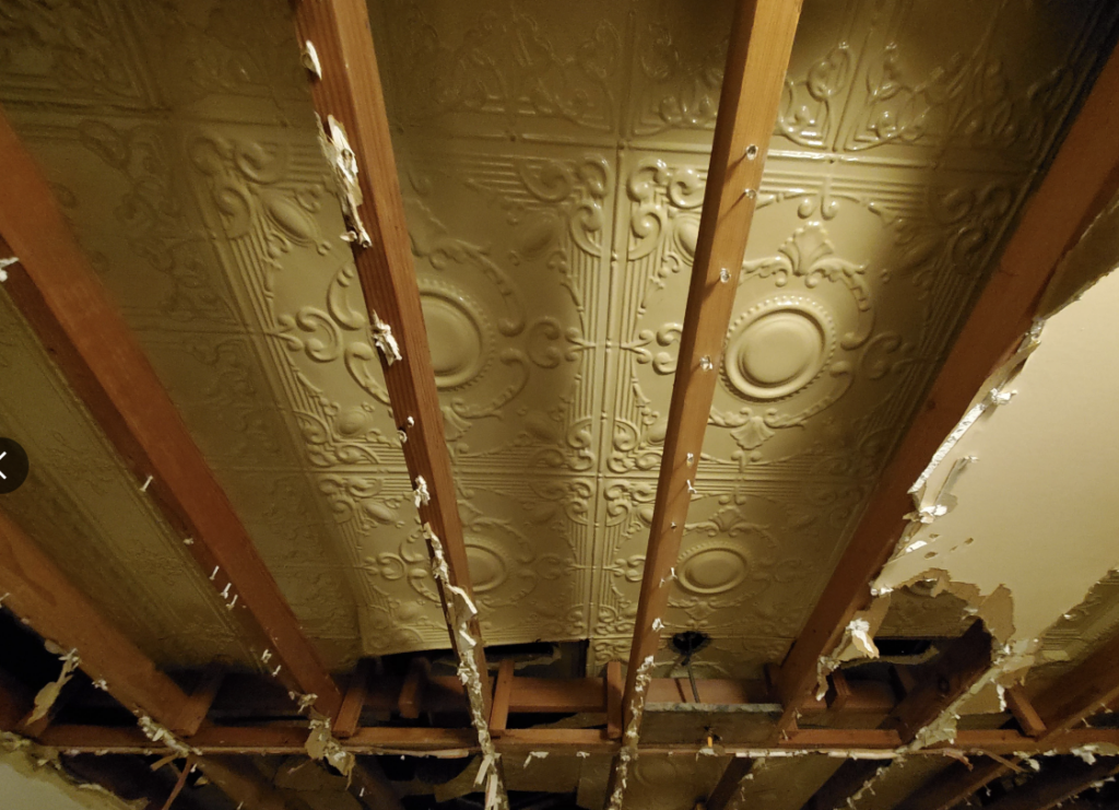 This image shows an ornate ceiling with an intricate pattern, partially exposed due to missing wooden beams. Pieces of debris and small bits of plaster hang off the remaining beams, revealing a view of the delicate design on the ceiling above.