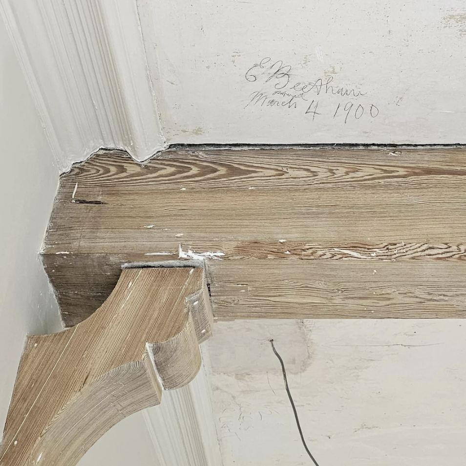A wooden beam in a corner with decorative molding, displaying the handwritten note "E. Bez Arani March 4 1900" on the adjacent wall. The beam and wall have an old, slightly worn appearance.