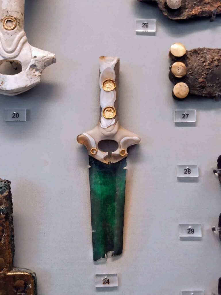 Photograph of a jade-hilted and green-bladed ceremonial dagger, displayed vertically in a museum exhibit. 