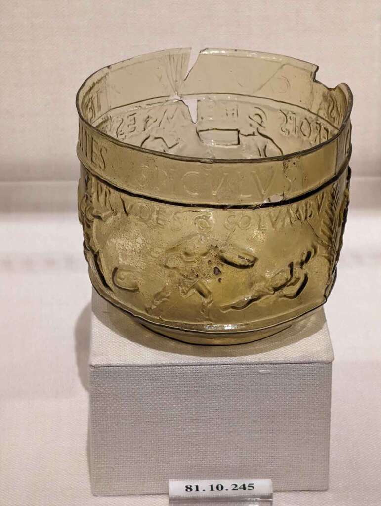 A partially broken transparent glass bowl displayed on a white pedestal. The bowl has an embossed design featuring horses and riders, along with some text near the rim. A small label on the pedestal reads "81.10.245".