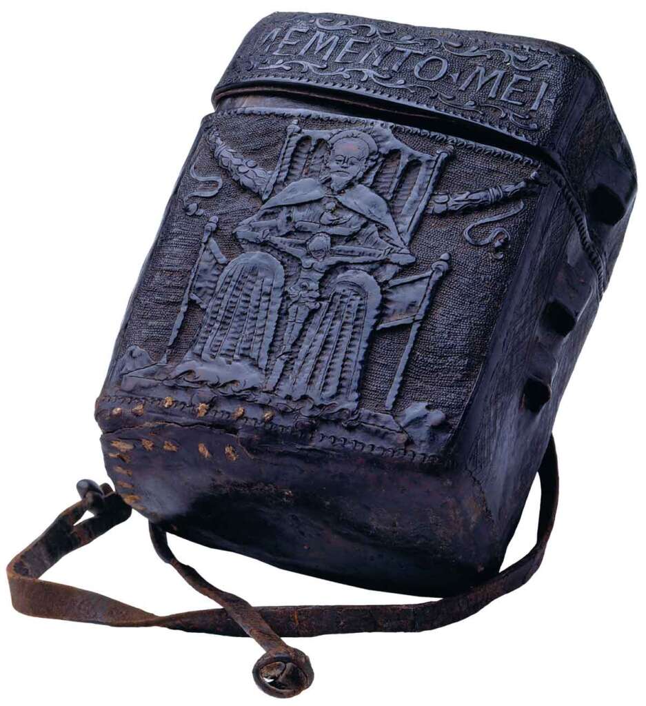 A dark leather bag with intricate carvings, including a figure seated on a throne, and Greek lettering at the top. The bag has a long strap attached, suggesting it is meant to be worn over the shoulder. The design appears ancient and detailed.