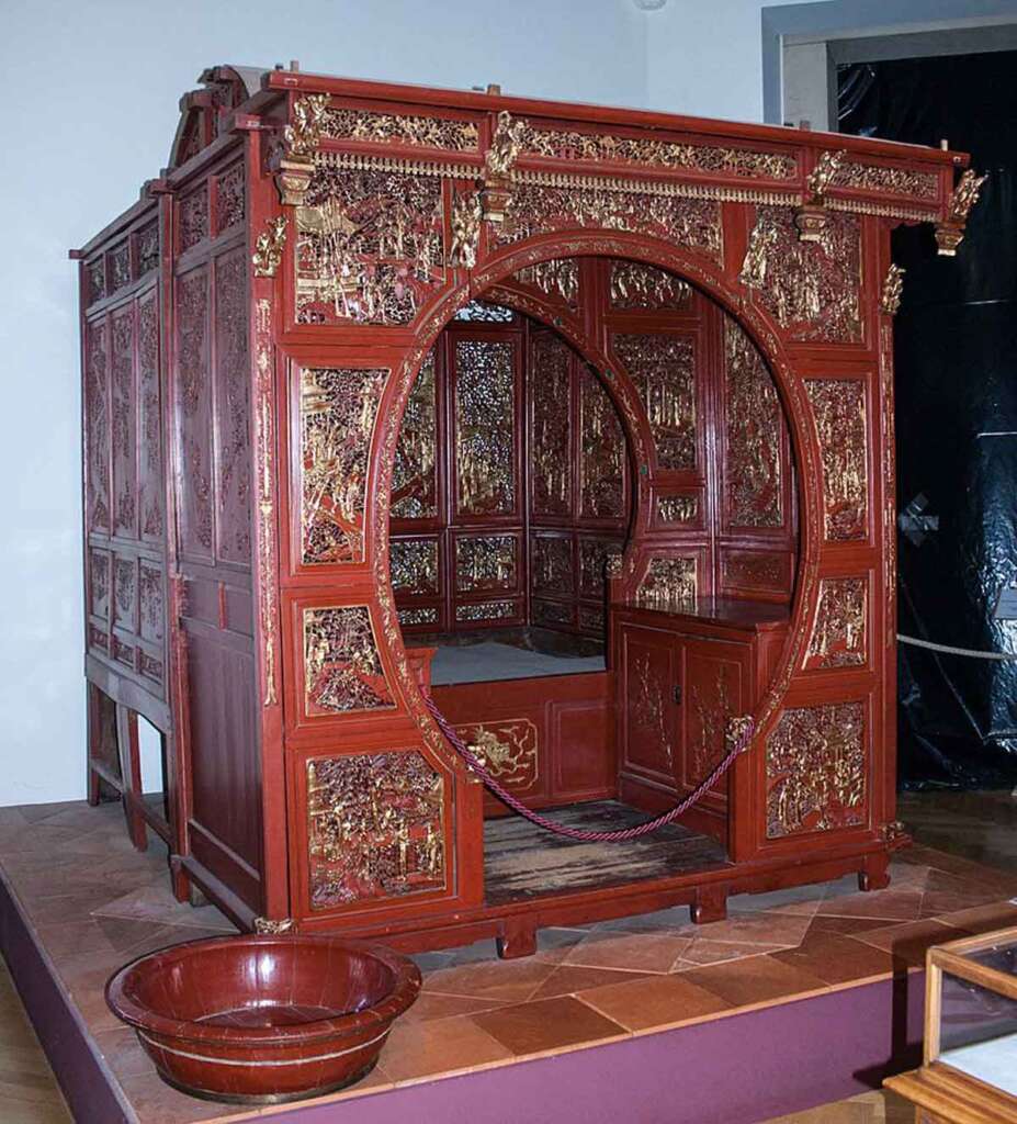 A richly adorned, traditional Chinese wedding bed with intricate red and gold carvings. The ornate wooden structure features a circular entrance with elaborate designs, set against a backdrop of detailed panels. A red bowl sits in front of the bed.