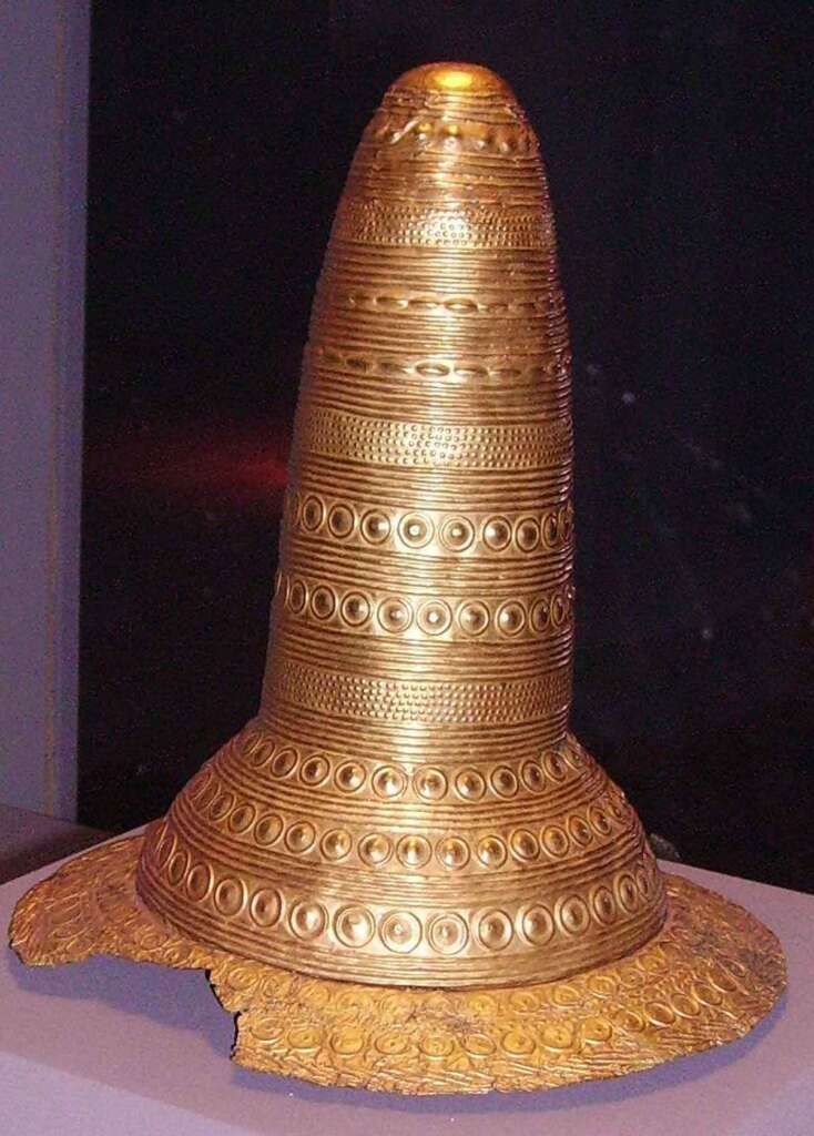 Tall, conical golden hat adorned with intricate circular patterns and bands displayed in a museum setting. The brim shows detailed designs with some signs of wear and damage.