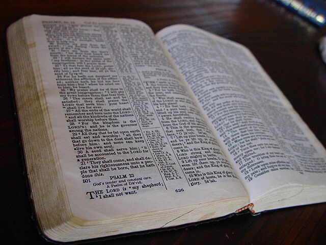 Open Bible lying on a dark wooden surface with visible text from the Book of Psalms. The Bible pages are slightly aged, with clear columns of text and verse numbers. The page on the right includes Psalms 22 and 23.