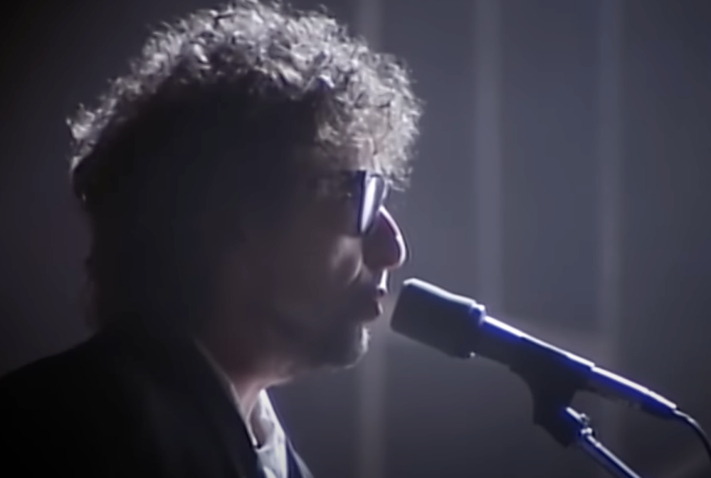 An image of Bob Dylan singing with sunglasses on. 