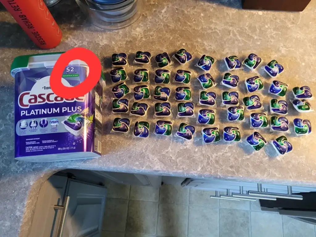 A plastic container of Cascade Platinum Plus dishwasher pods is open on a kitchen counter. Beside it, there are 47 individual dishwasher pods arranged neatly in rows. The container is labeled to contain 52 pods.
