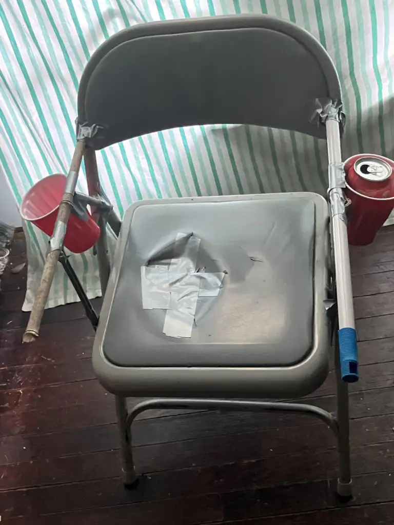 A gray folding chair with armrests made of a wooden pole and a metal rod, both attached with duct tape. A red plastic cup is taped to each side as cup holders, and the chair seat is covered with a silver duct tape X. The background has a green-striped fabric.