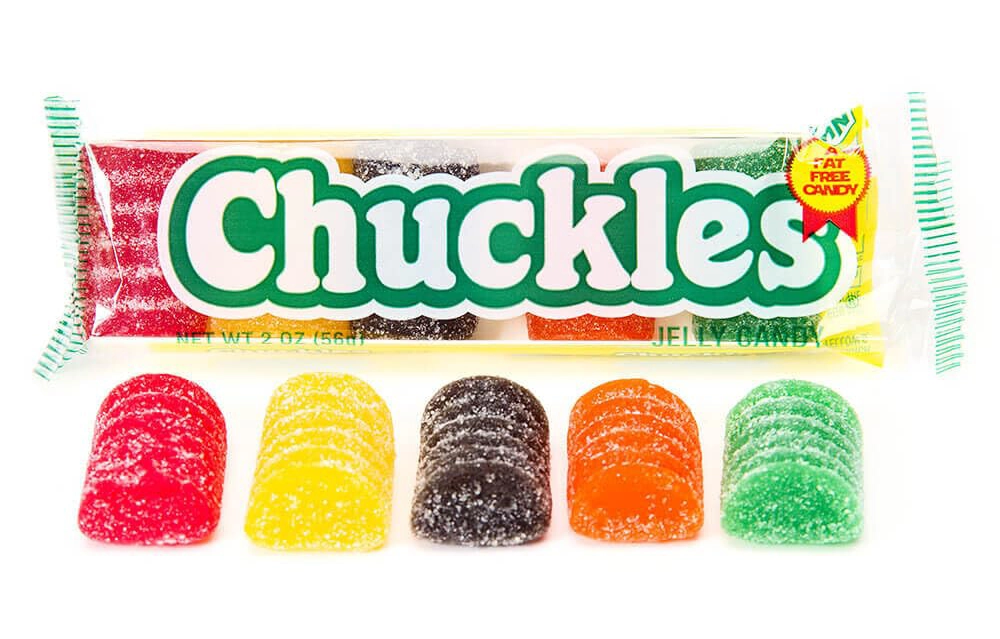 The image shows a package of Chuckles jelly candies. Five sugar-coated candies in different colors (red, yellow, green, orange, and black) are lined up in front of the package. The candies are displayed neatly in a transparent wrapper with the brand name "Chuckles.