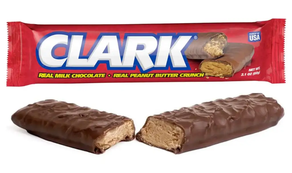 A Clark candy bar wrapper with "CLARK" in bold white letters on a red background. The text says "Real Milk Chocolate" and "Real Peanut Butter Crunch." Below, an unwrapped bar is split in half, revealing a creamy peanut butter core inside a chocolate coating.