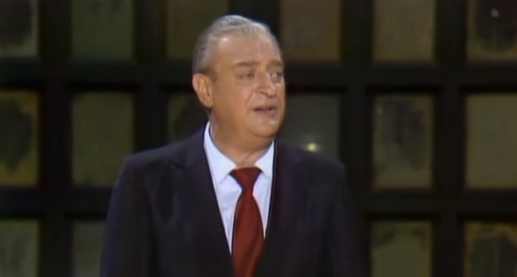 A man with gray hair, wearing a dark suit, white shirt, and red tie, is standing in front of a grid of darkened windows. He appears to be in the middle of speaking or performing. The background is dimly lit.