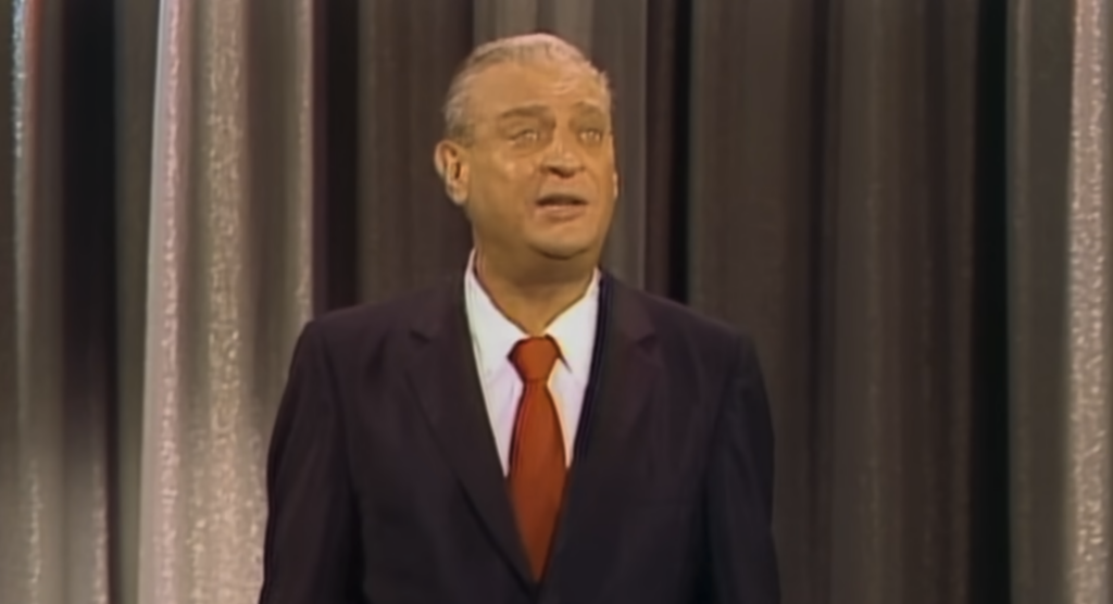 A man in a dark suit, white shirt, and red tie is standing in front of a curtain and looking upward. He has light-colored hair combed back. The background consists of vertically hanging, two-tone curtains.