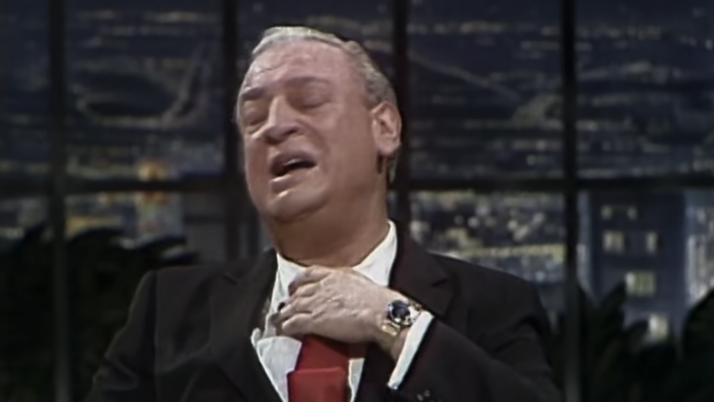 A man with light-colored hair wearing a dark suit and red tie appears to be adjusting his tie while making an expressive face. The background is a blurred interior setting with lights, suggesting a nighttime talk show or studio environment.