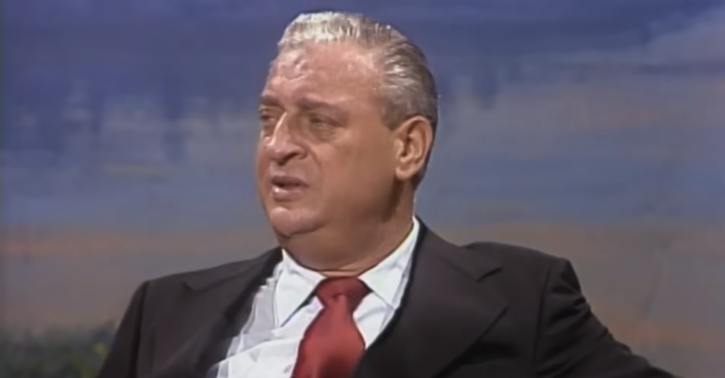 A person with white slicked-back hair, wearing a dark suit, white shirt, and red tie, is seen sitting against a blurred background. The individual appears engaged in conversation, with a neutral to slightly concerned facial expression.