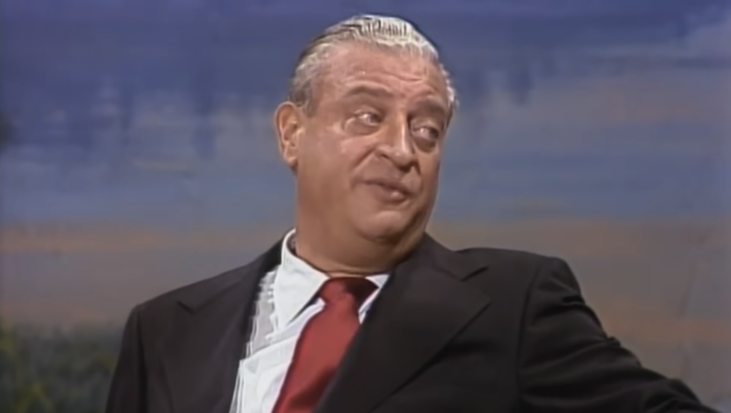 A man with slicked-back gray hair, wearing a black suit, white dress shirt, and red tie, is seen from the waist up. He is sitting and leaning slightly to one side, looking off to his left. The background is somewhat abstract with blue and earthy tones.